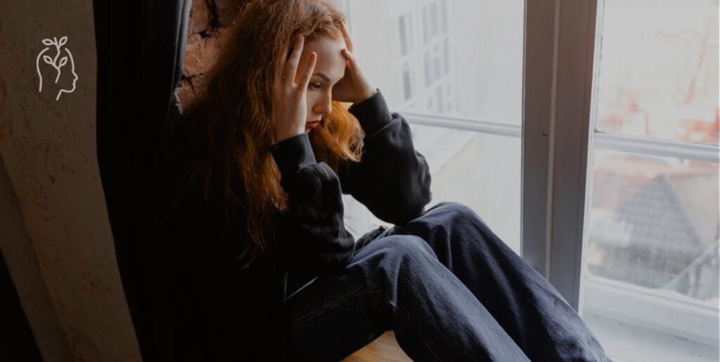 Jeune femme rousse assise à sa fenêtre qui se prend la tête dans ses mains