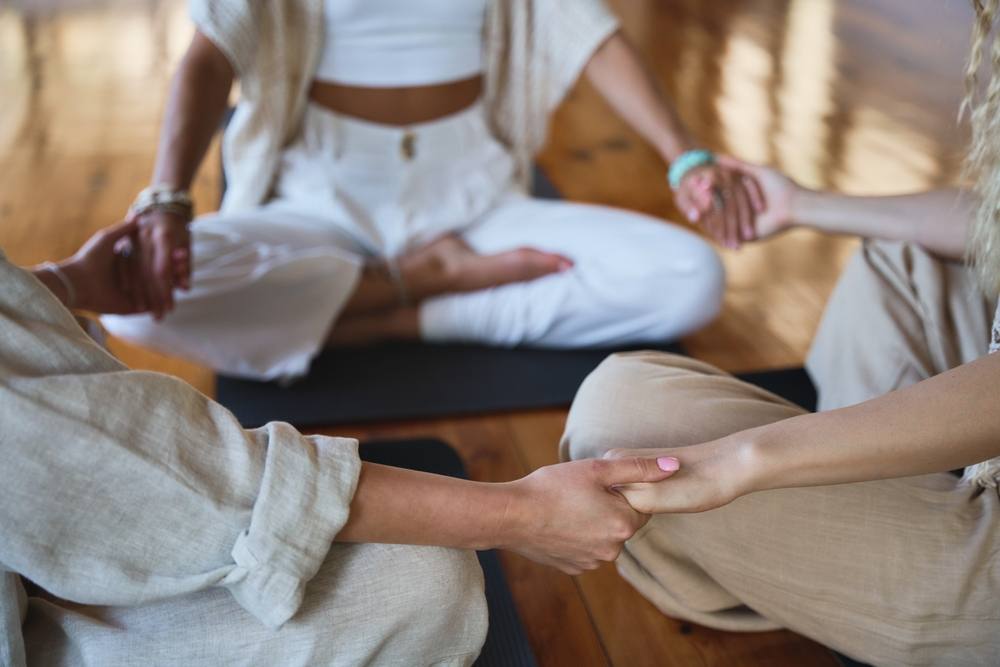 trois personnes assises se tenant par les mains pour faire une méditation collective