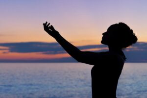 jeune femme au coucher du soleil sur une plage en train de pratiquer la méditation en levant ses mains au ciel